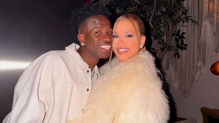 Homem e mulher sorriem enquanto posam juntos para foto em ambiente interno de evento, com iluminação suave e decoração ao fundo.