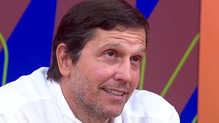 Homem de camisa clara sorri durante participação em programa de TV com cenário colorido ao fundo