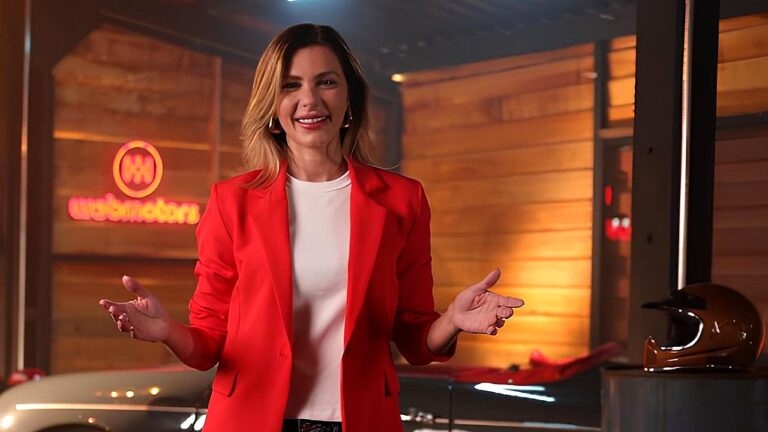 Apresentadora de blazer vermelho fala em estúdio com logotipo do Webmotors ao fundo