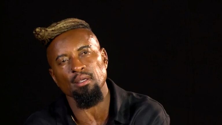 Homem com barba e cavanhaque, usando brinco e um penteado preso no topo da cabeça (tipo dread ou coque), falando ou sendo entrevistado diante de um fundo escuro.