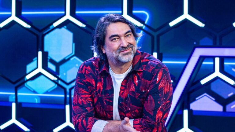 Zeca Camargo inclina o corpo e sorri no cenário iluminado com painéis geométricos do programa 1001 Perguntas da Band