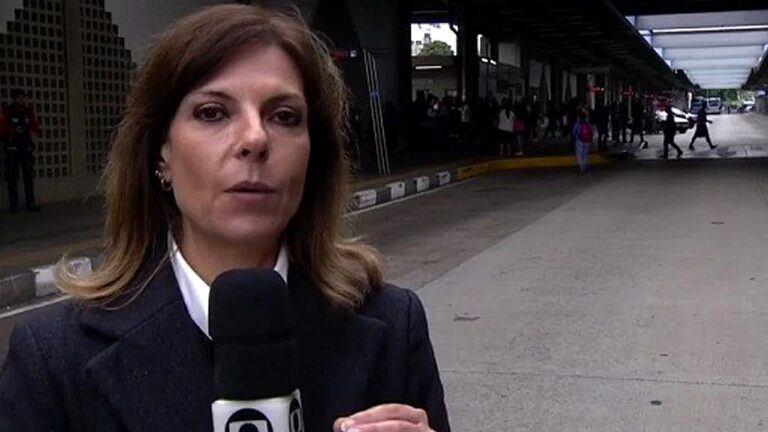 Repórter segura microfone com logotipo da Globo durante entrada ao vivo em frente a prédio público com movimentação ao fundo