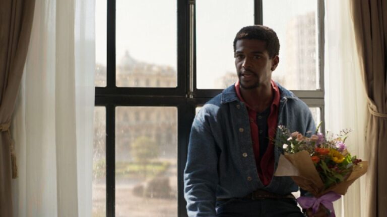 Homem segura buquê de flores e olha atento diante de janela em cena de série ou filme