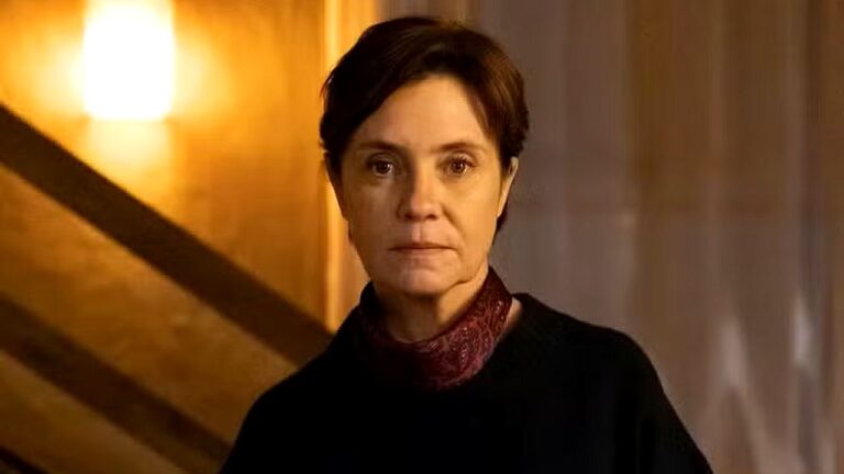 Mulher com expressão séria observa à frente em cena dramática com iluminação quente em ambiente interno