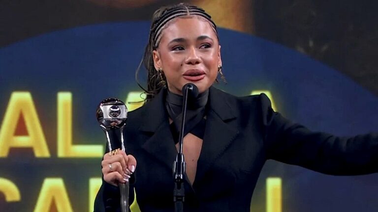 Atriz recebe prêmio e discursa ao microfone em cerimônia com troféu nas mãos