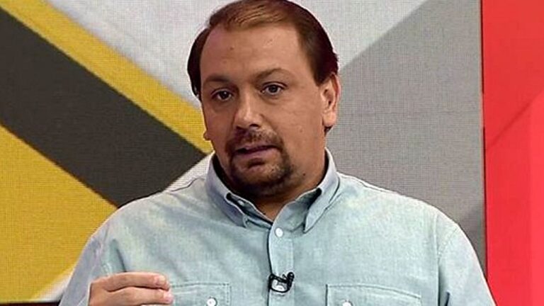 Homem com microfone de lapela fala em estúdio de TV diante de painel com gráfico ao fundo