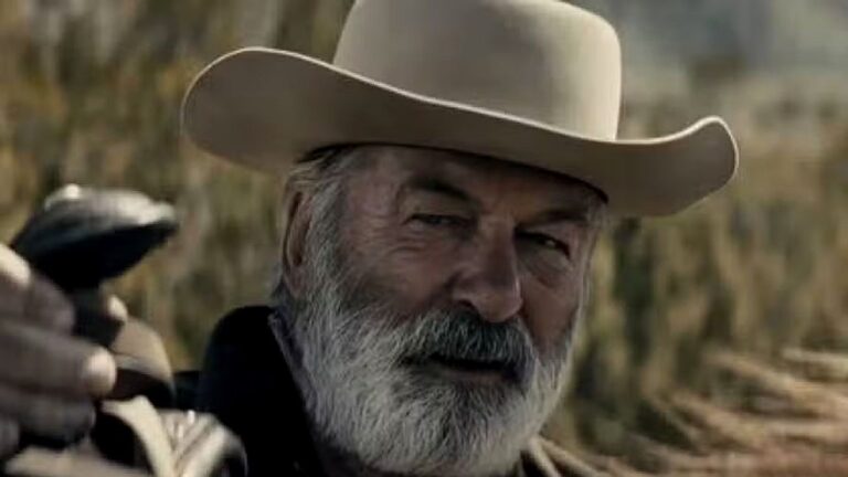 Homem com chapéu de cowboy e barba grisalha aponta arma em cena ambientada no campo