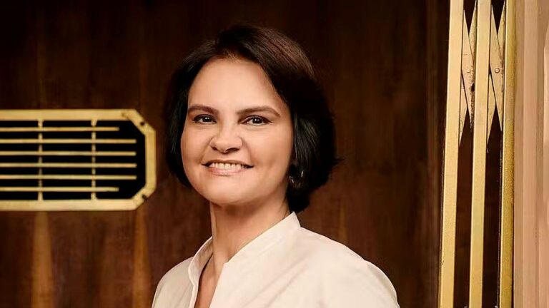 Mulher sorri diante de porta de madeira em cena de novela ambientada em interior elegante