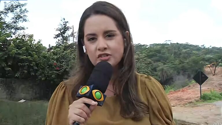 Alice Ribeiro, repórter da Band, segura microfone durante entrada ao vivo em área com vegetação e estrada de terra ao fundo.