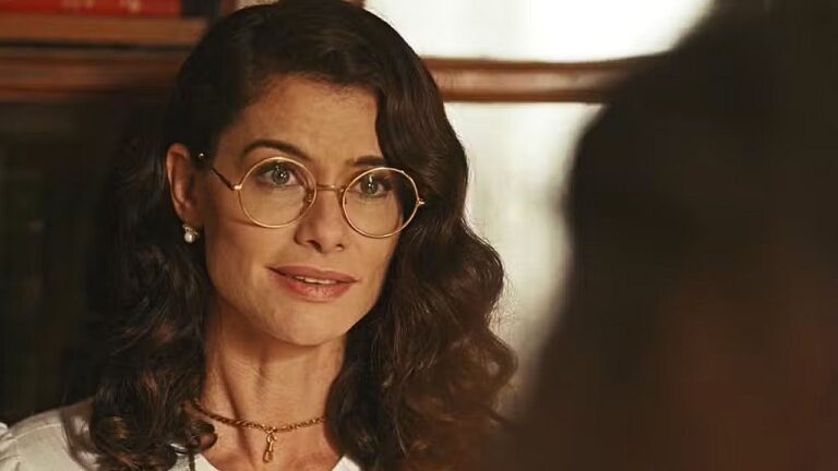 Mulher de óculos redondos e cabelo ondulado conversa com alguém em cena de novela ambientada em interior