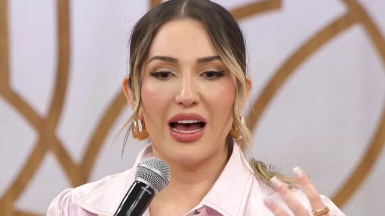 Mulher fala ao microfone durante entrevista em estúdio usando blusa rosa e brincos dourados