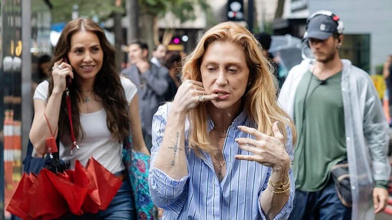 Mulher faz gesto de telefone com a mão enquanto caminha na rua acompanhada por outra mulher segurando guarda-chuva