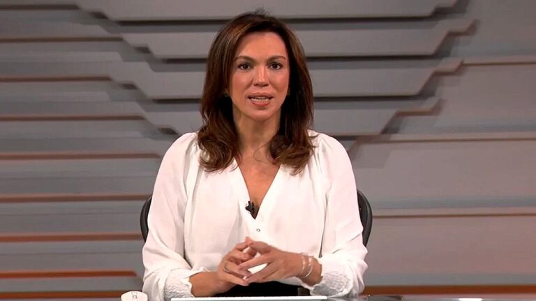 Apresentadora de telejornal veste blusa branca e fala ao público em estúdio com cenário geométrico ao fundo