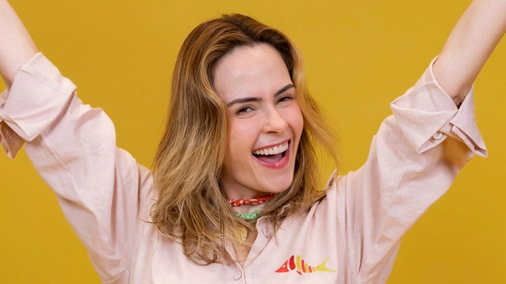 Ana Paula Renault sorrindo com braços erguidos usando camisa clara diante de fundo amarelo em foto promocional