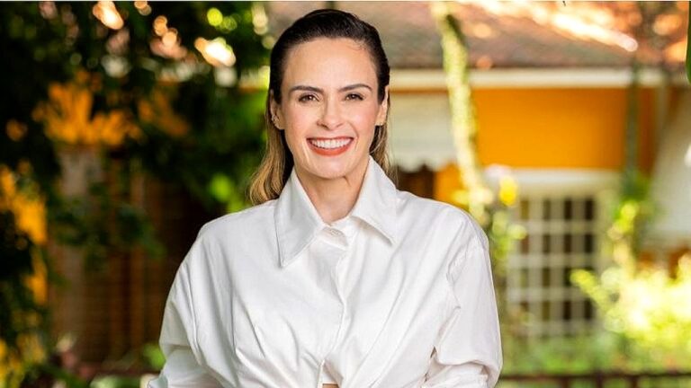 Mulher sorri usando camisa branca em área externa com jardim e casa ao fundo em foto promocional