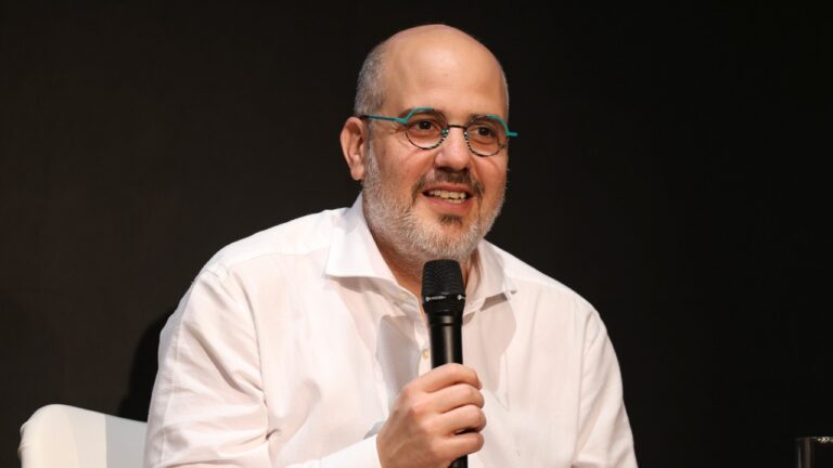 André Basbaum fala ao microfone durante evento em palco com fundo escuro