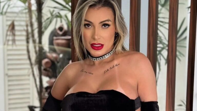 Andressa Urach posa com vestido preto e batom vermelho em ambiente interno com plantas e portas de madeira ao fundo