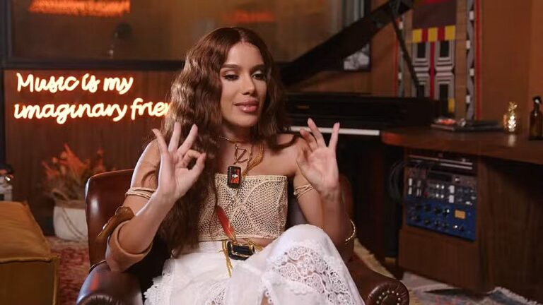 Cantora sentada em estúdio faz gestos com as mãos durante entrevista com piano e letreiro ao fundo