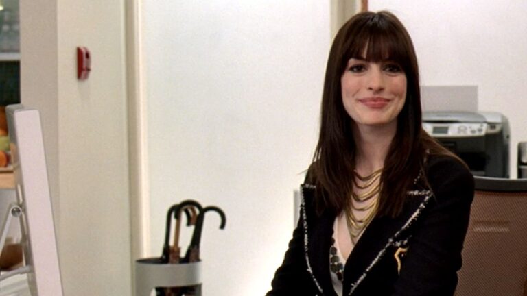 Mulher de cabelo castanho com franja sorri sentada em escritório com computador e guarda-chuvas ao fundo