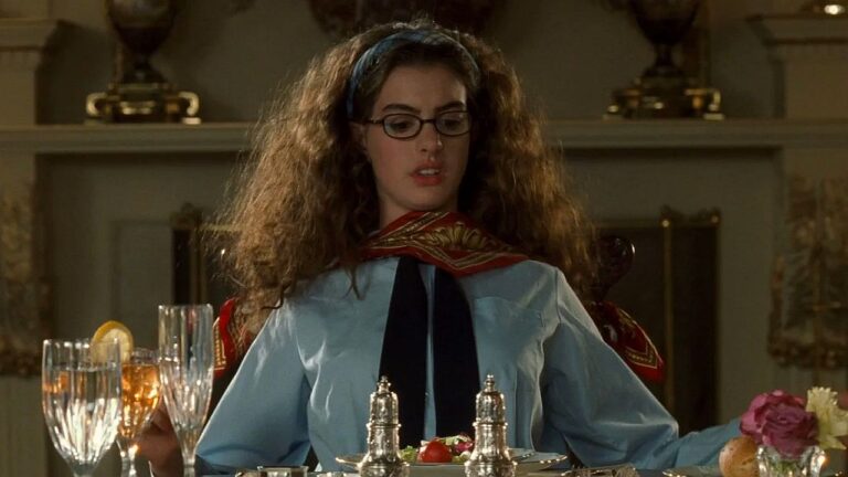 Mulher com cabelo volumoso e óculos sentada à mesa posta em cena de jantar formal em filme de comédia romântica