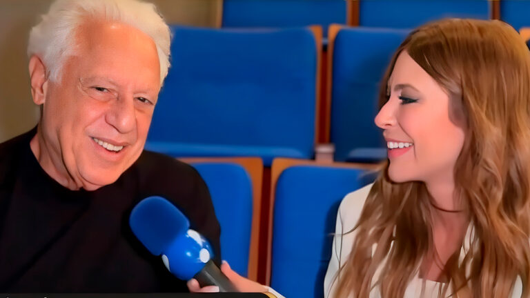 Antonio Fagundes fala ao microfone azul enquanto Naty Curvelo sorri durante entrevista em auditório com poltronas azuis ao fundo