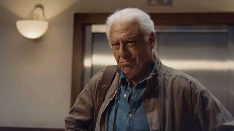 Homem idoso de cabelos brancos com mochila observa com expressão séria em corredor interno próximo a elevador