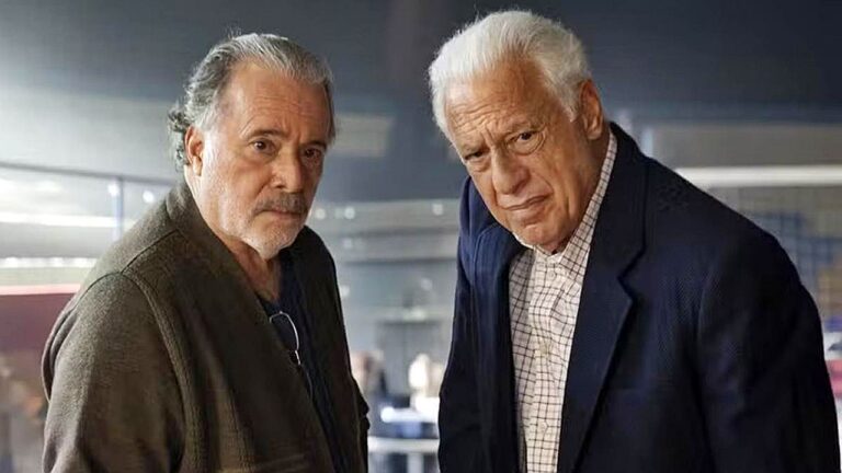 Dois homens idosos com expressão séria conversam em ambiente interno em cena de série ou filme