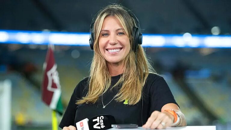 Repórter esportiva com fone de ouvido segura microfone da Cazé TV durante transmissão em estádio ao fundo