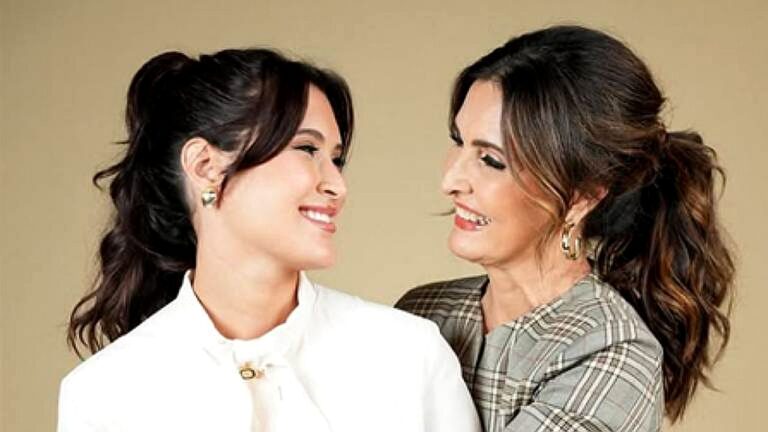 Duas mulheres sorrindo se abraçam e trocam olhares em ensaio fotográfico com fundo neutro