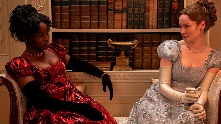 Duas mulheres em vestidos de época conversam sentadas em sala com estante de livros ao fundo