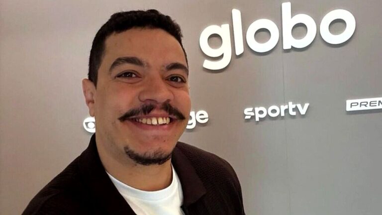 Homem sorri em selfie diante de painel com logotipos da Globo e SporTV em ambiente corporativo