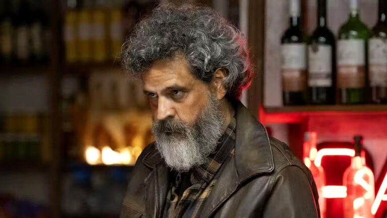 Homem de barba grisalha e casaco de couro encara sério em bar com garrafas ao fundo