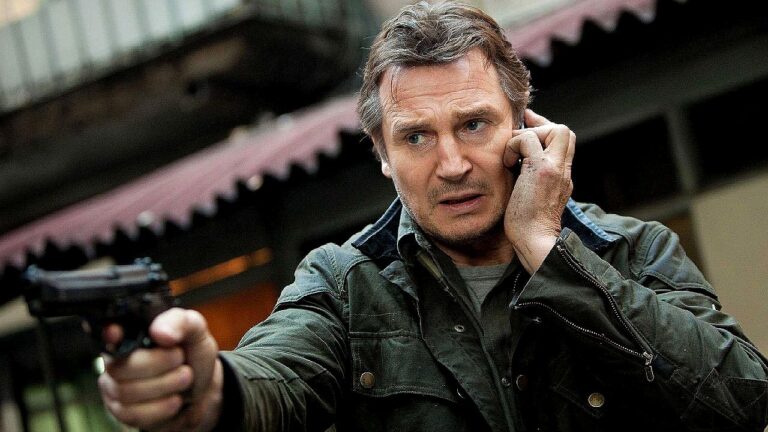 Liam Neeson fala ao celular enquanto aponta arma em cena de filme de ação