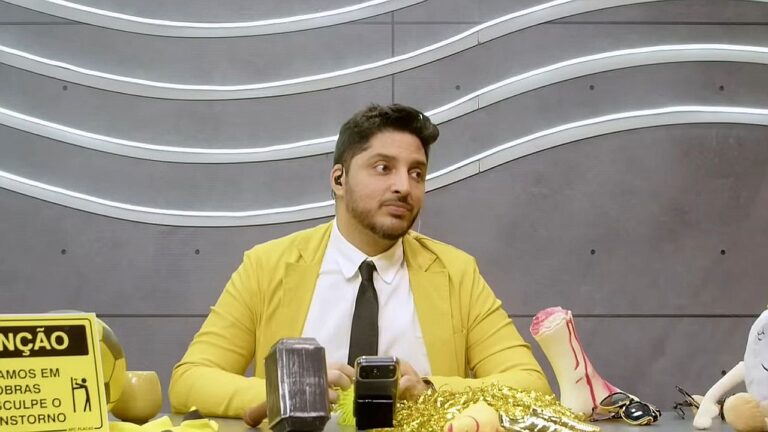 Homem de blazer amarelo sentado em bancada de programa com objetos variados e celular sobre a mesa