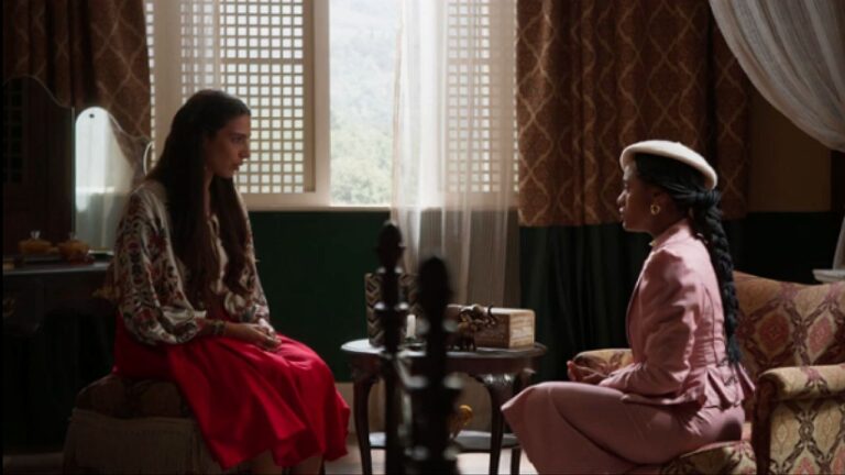 Duas mulheres sentadas frente a frente conversam em sala com decoração clássica e luz natural entrando pela janela