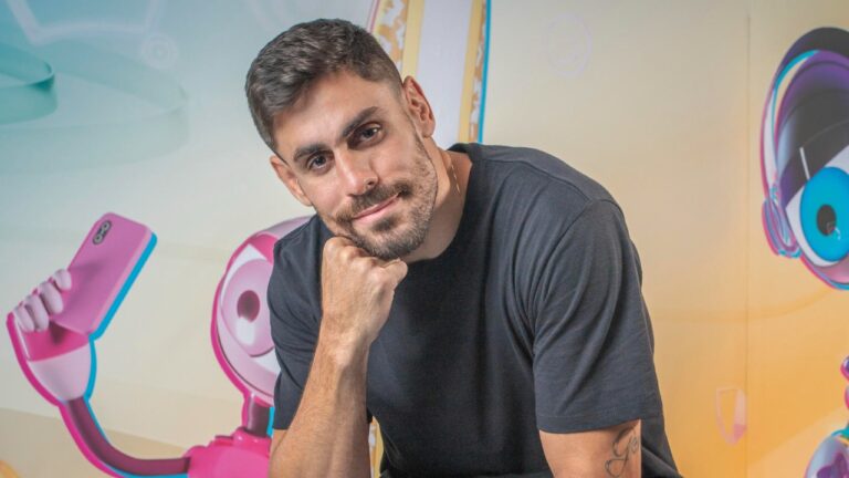 Cara de Sapato apoia o queixo na mão e olha para a câmera em cenário colorido com ilustrações de celular e fones ao fundo