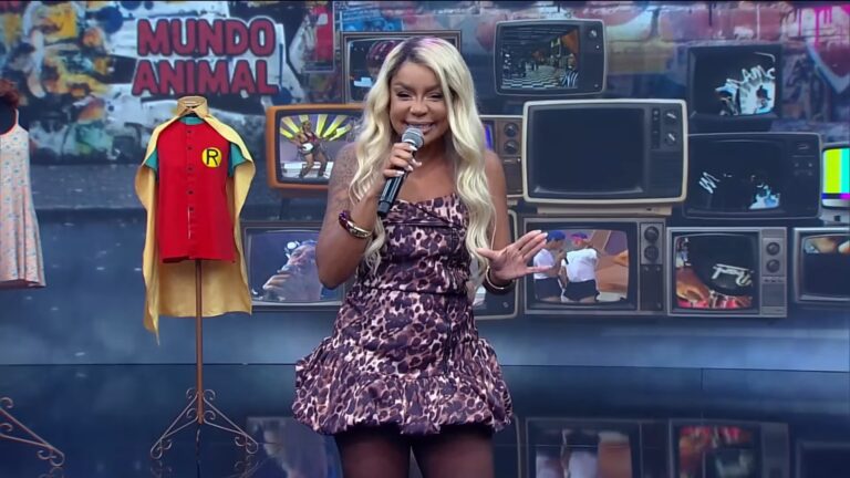 Cariúcha, apresentadora com um vestido animal print, segura microfone no palco do programa Superpop, da RedeTV!, com TVs ao fundo.