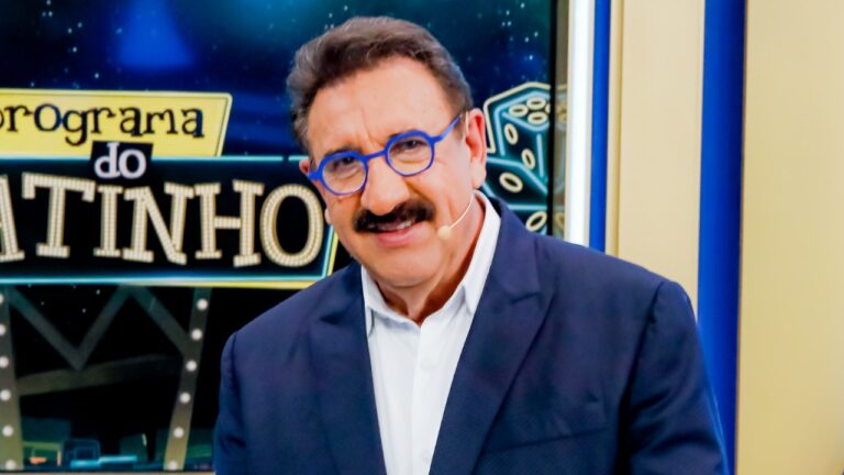 Ratinho sorri usando óculos azuis e microfone de cabeça no estúdio do Programa do Ratinho com logo ao fundo