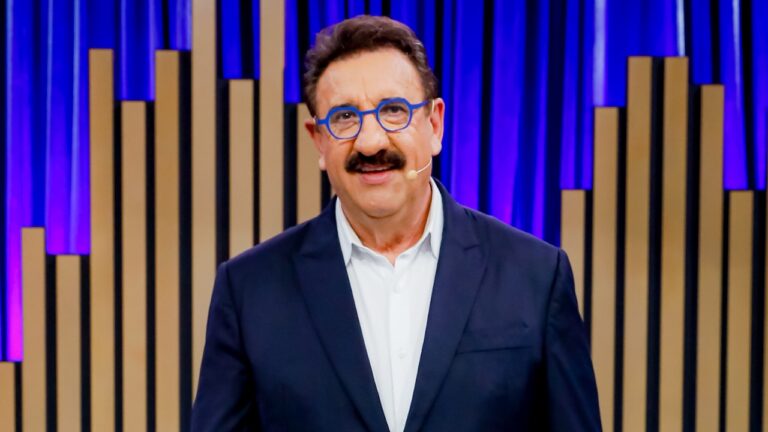 Ratinho apresenta programa em estúdio com fundo de painéis verticais iluminados usando terno escuro óculos azuis e microfone headset