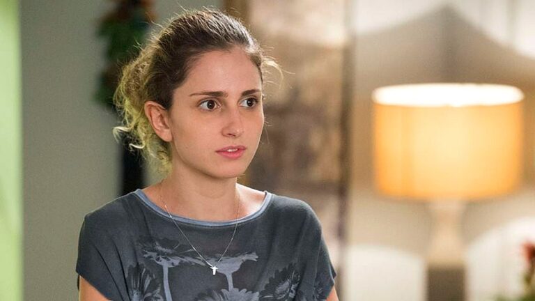 Jovem com expressão preocupada usa camiseta cinza e colar com cruz em cena de novela dentro de casa