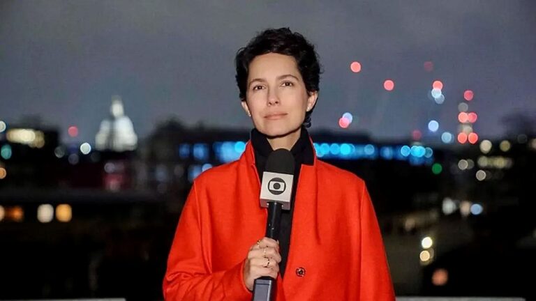 Repórter segura microfone da TV Globo durante entrada ao vivo à noite com cidade iluminada ao fundo