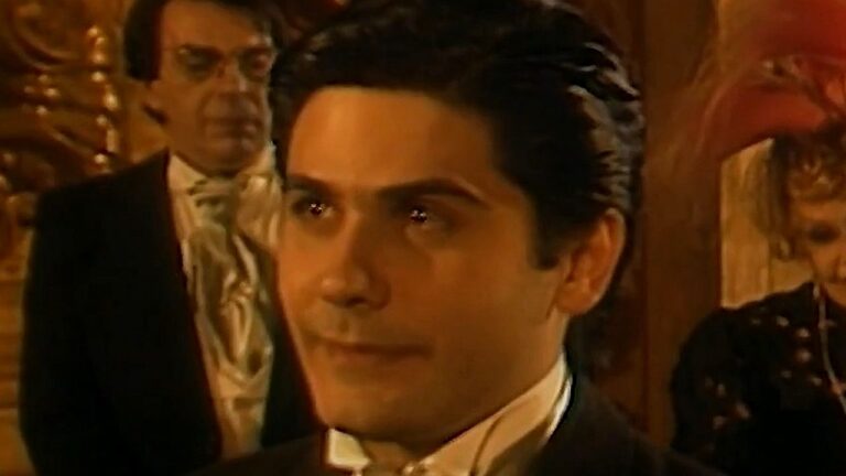 Homem de terno com expressão séria em primeiro plano enquanto convidados aparecem ao fundo em cena de novela de época