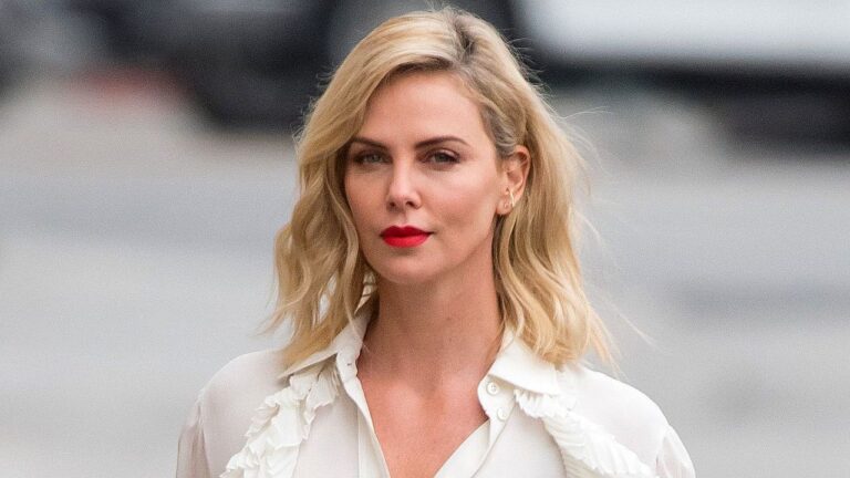 Mulher loira com batom vermelho e blusa branca caminha na rua com expressão séria