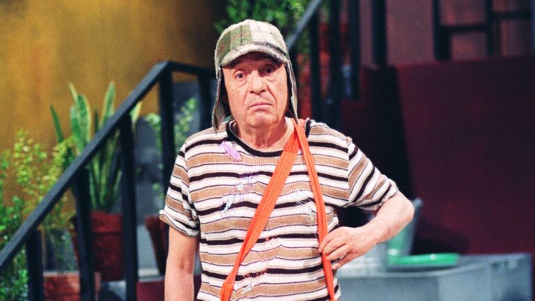 Chaves (Roberto Gómez Bolaños) com chapéu e suspensório laranja em cenário de vila com escada e plantas ao fundo