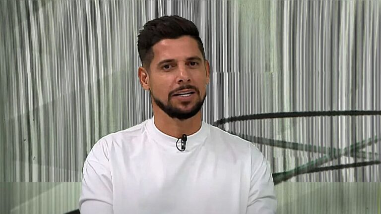 Apresentador de camiseta branca fala em estúdio de TV com microfone de lapela preso à roupa