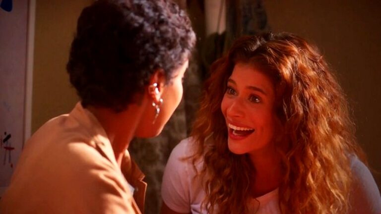 Mulher sorri animada enquanto conversa com outra em cena de novela com iluminação quente em ambiente interno