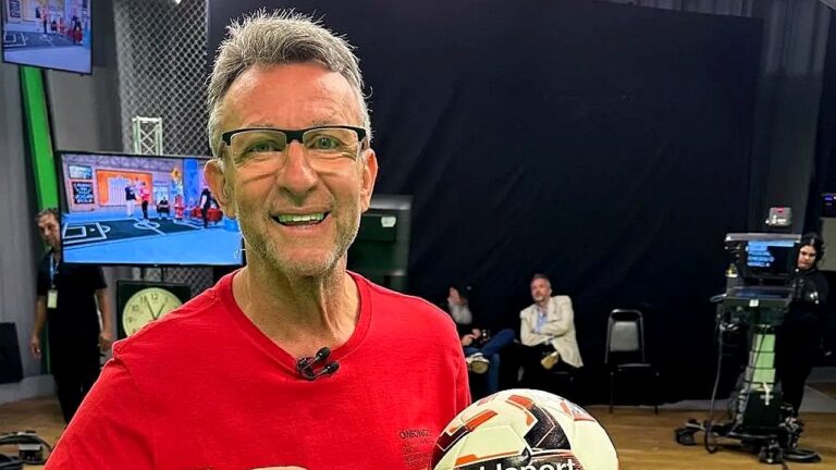 Apresentador esportivo segura bola de futebol em estúdio de TV com câmeras e equipe ao fundo