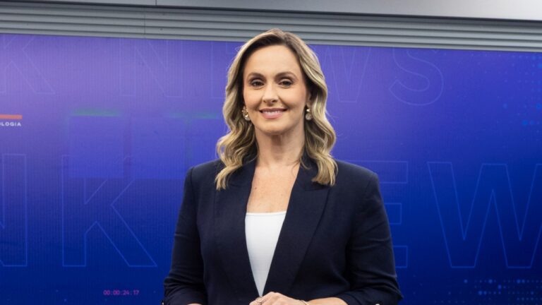 Daysa Belini sorri em estúdio da Record News com telão azul ao fundo exibindo identidade visual do telejornal