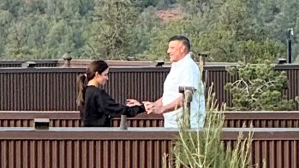 Homem e mulher seguram as mãos em área externa com cerca de madeira e vegetação ao fundo