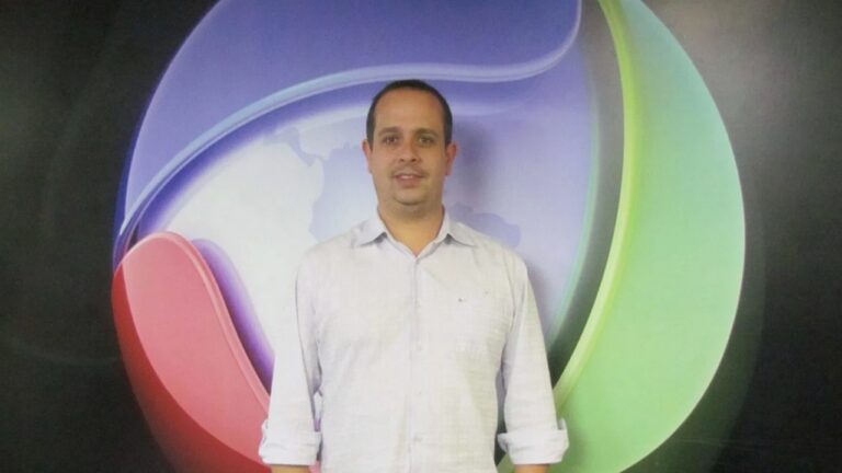 Eduardo Caruso, de camisa clara posa, diante de logotipo colorido da Record em fundo escuro.